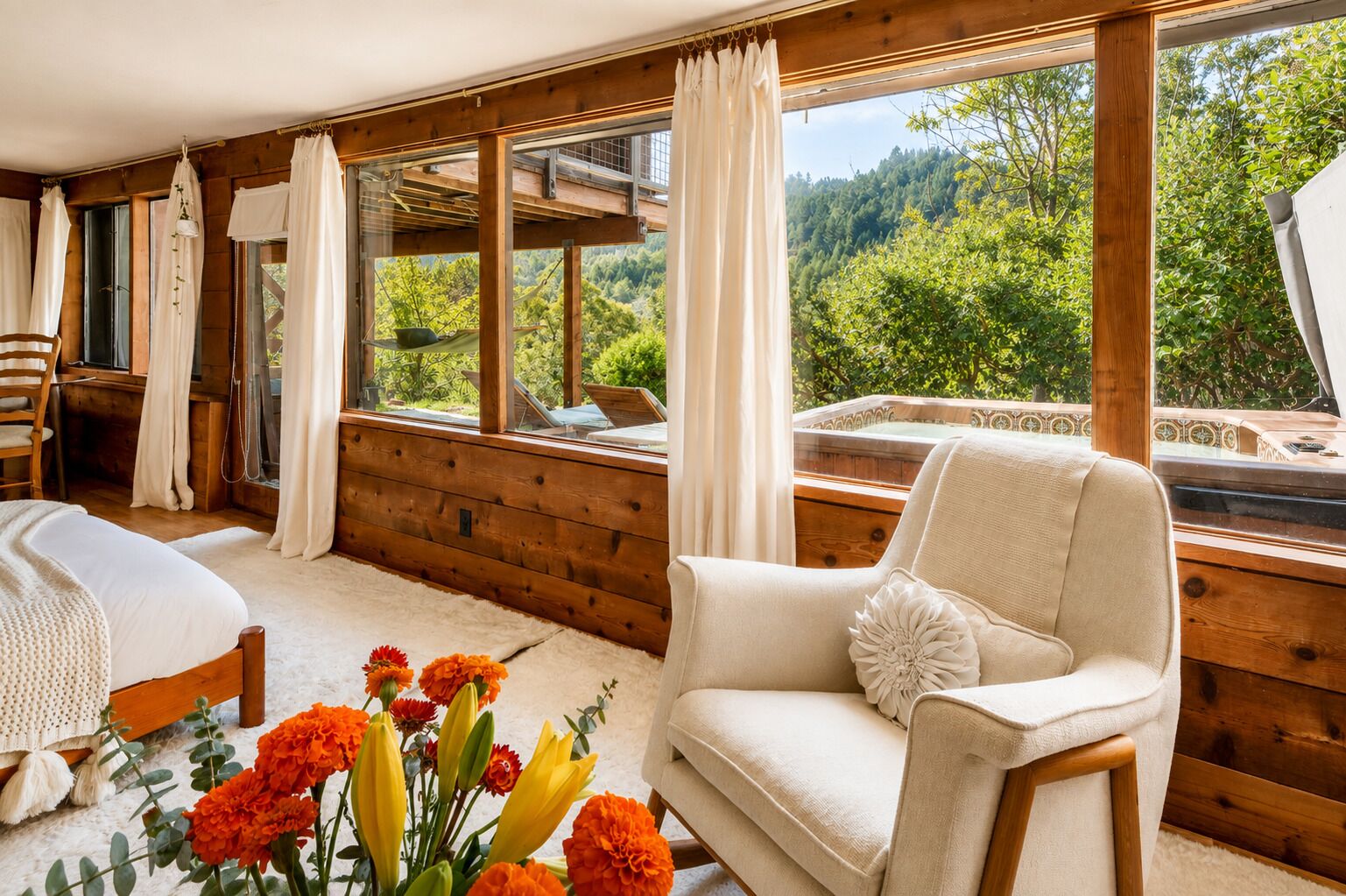 Ein gemütliches Zimmer mit Holzvertäfelung, einem Bett, einem weißen Sessel und Blick auf einen Wald durch große Fenster.
