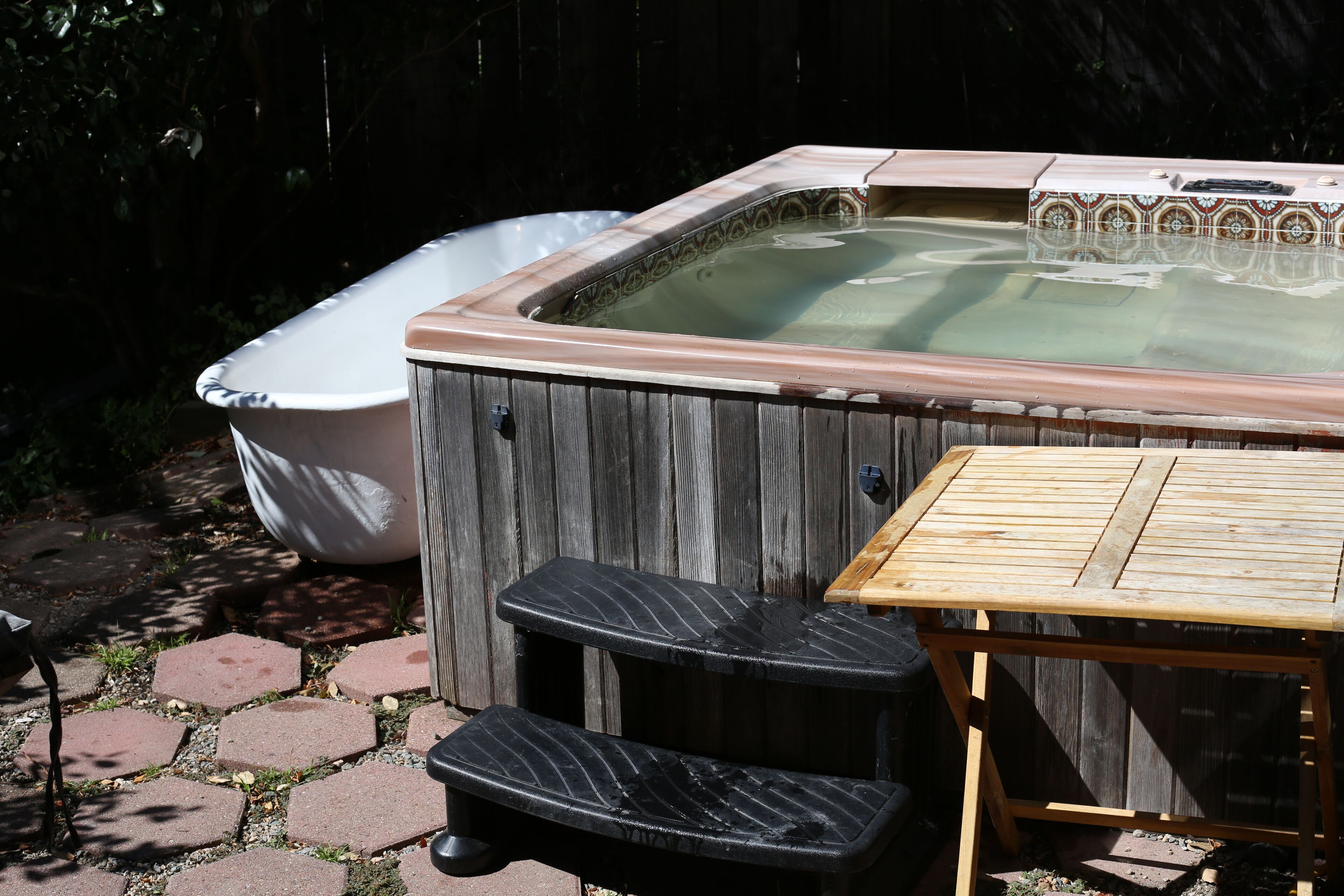 A hot tub with a wooden deck and steps, surrounded by a wooden fence and a brick pathway.
