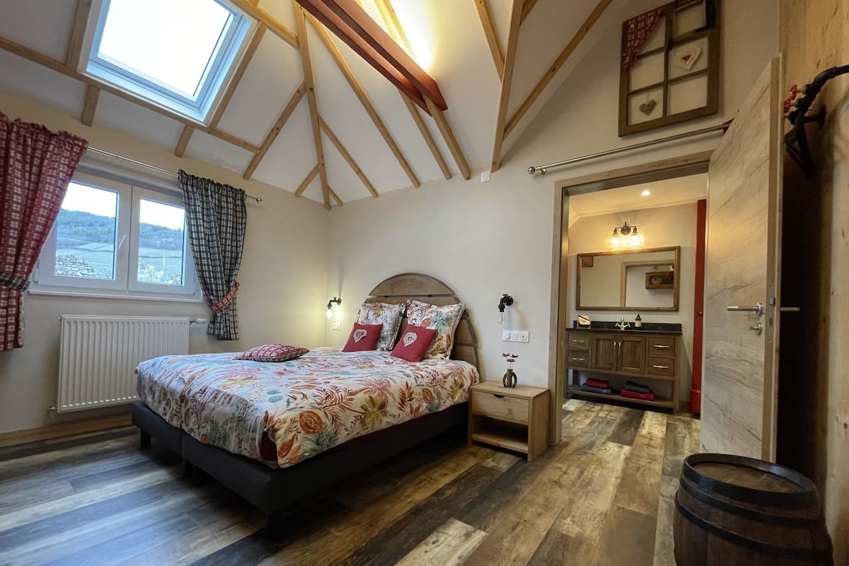 Un dormitorio con cama, suelo de madera, tragaluz, ventana, mesita de noche, espejo y puerta.