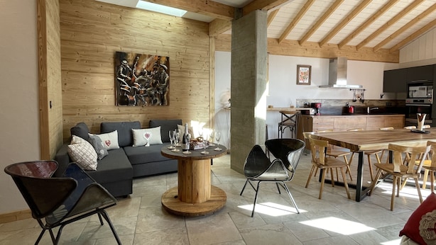 Een ruime woonkamer met houten plafond, een eettafel met stoelen, een bank met kussens en een salontafel.