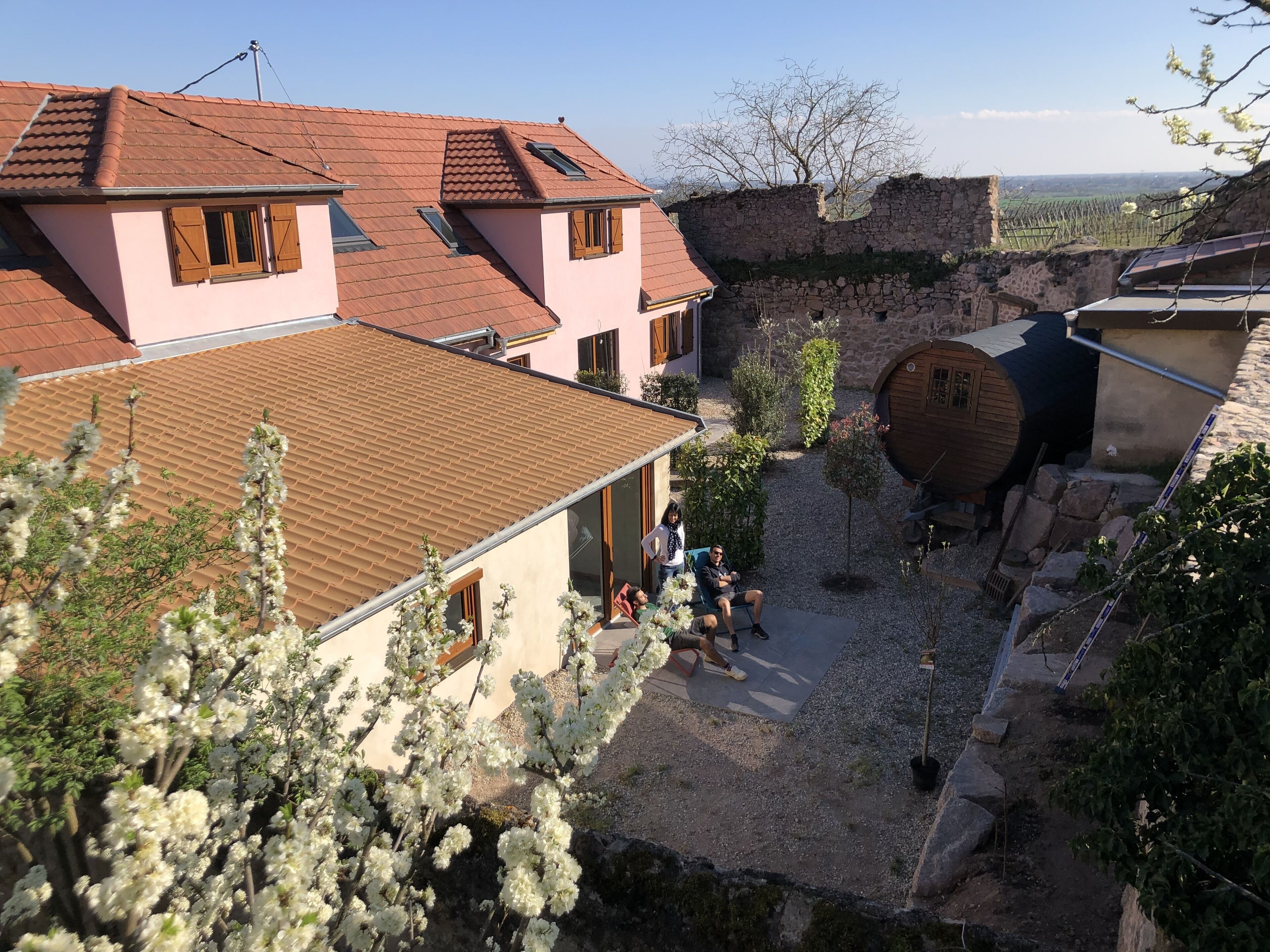 Une cour bordée de murs en pierre, un tonneau en bois et un bâtiment surmonté d’un toit en tuiles.