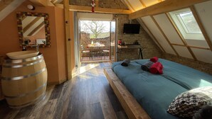 Slaapkamer met schuine plafond, houten balken, een stenen muur, een bed met kussens, een houten vat en een raam.