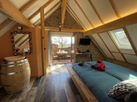 Un dormitorio con techo inclinado, vigas de madera, pared de piedra, cama con almohadas, un barril de madera y una ventana.
