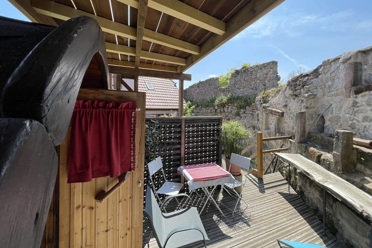 Una terraza de madera con una mesa y sillas, una cortina roja y un muro de piedra al fondo.