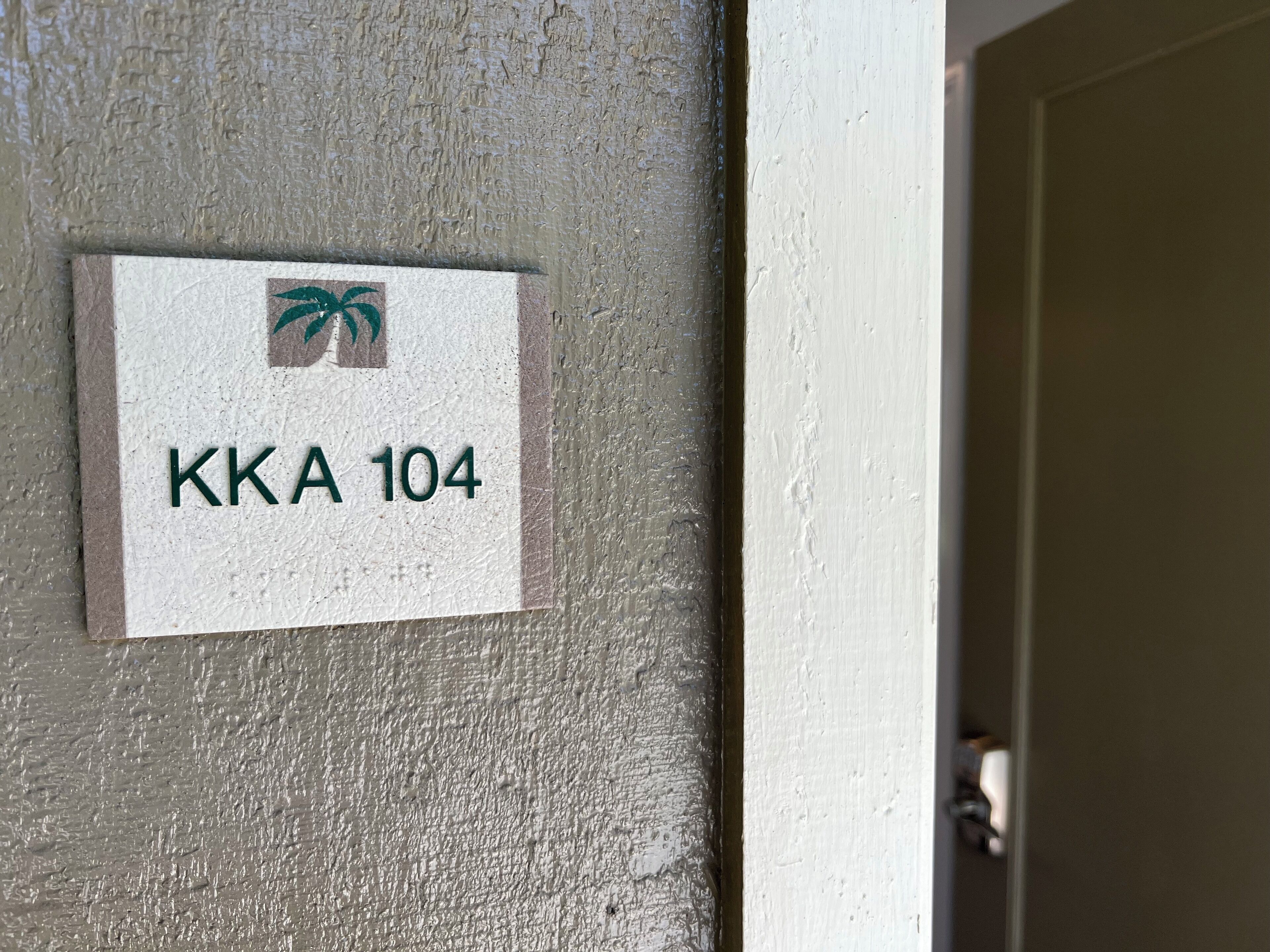 Een bord met 'KKA 104' en een palmboom logo op een gestructureerde muur.