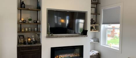 Una sala de estar con un televisor de pantalla plana sobre la chimenea, estantes de madera con objetos decorativos y un ventanal con persianas.