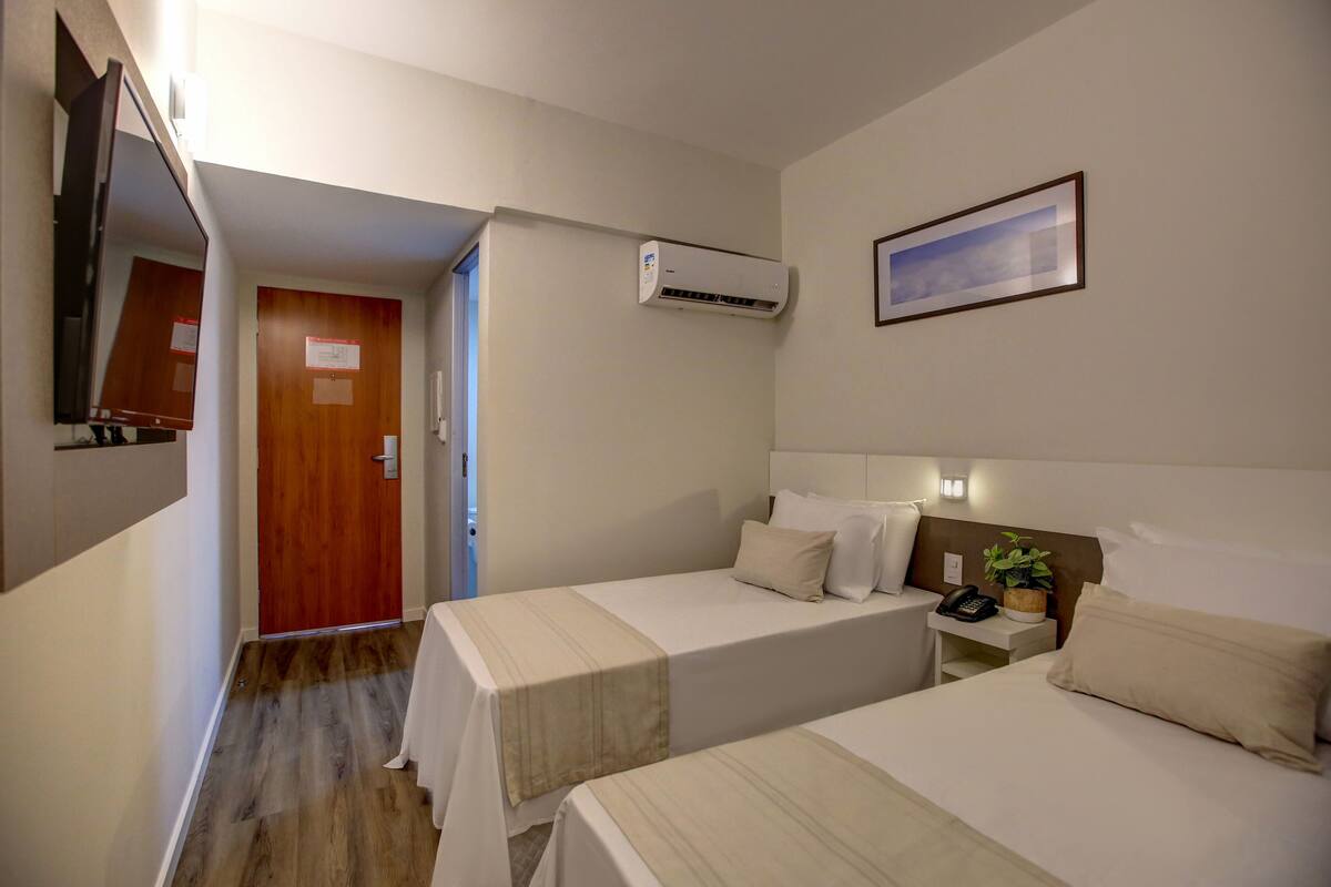 Quarto de hotel com duas camas, uma mesa de cabeceira com telefone e uma planta, um ar condicionado, um quadro na parede e uma porta de madeira.