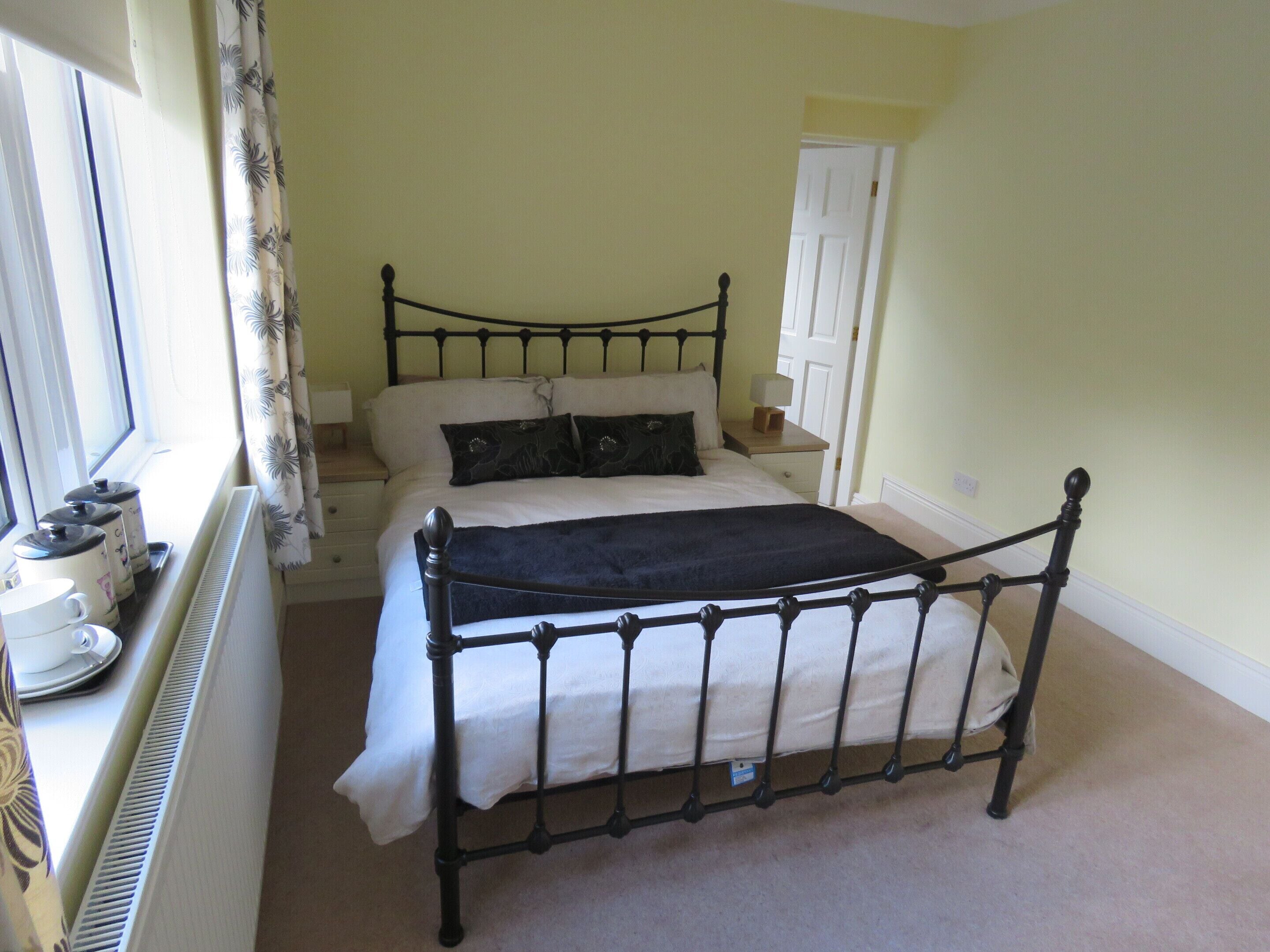 A neatly made bed with a black metal frame, a white duvet, and two dark pillows. There is a bedside table with a lamp and a small wooden box. A window with curtains is visible to the left.