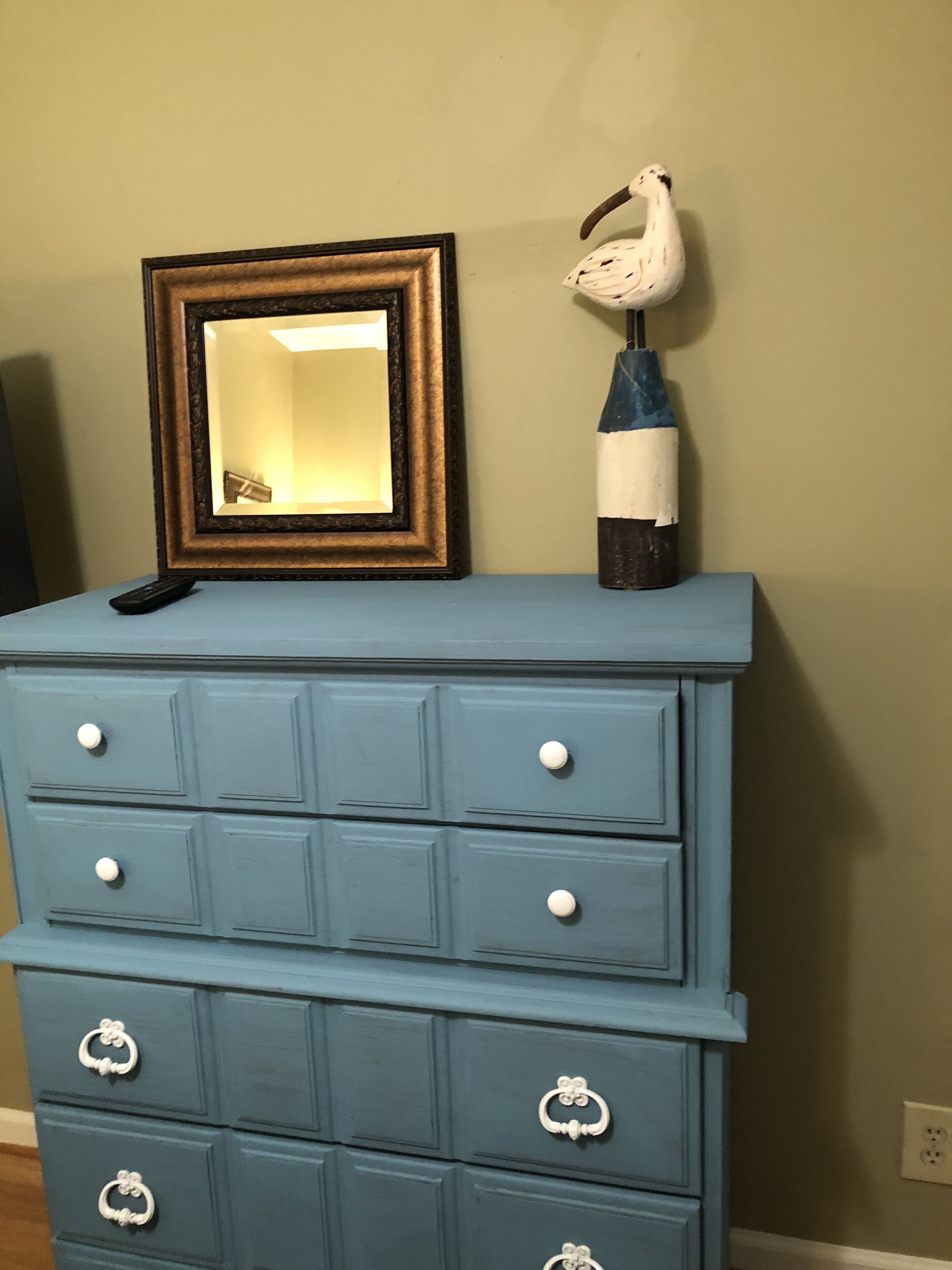 Une commode bleue avec des tiroirs et des boutons, un miroir et un vase décoratif posé sur le dessus.