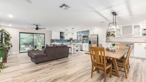 Una cocina moderna con armarios blancos, una zona de comedor con sillas de madera y un sofá gris en una sala de estar con un ventanal grande.