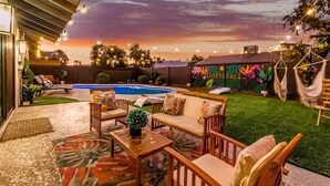 Zona de piscina con sillones reclinables y un sofá, rodeada de follaje y vistas al atardecer.