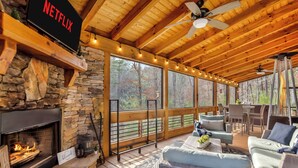 A cozy cabin interior with a stone fireplace, a Netflix sign, wooden ceiling beams, and a deck with outdoor furniture. - Cozy Blue Ridge Escape •Hot Tub•Fireplace-2 acres with covered porch/Fireplace (Mineral Bluff)