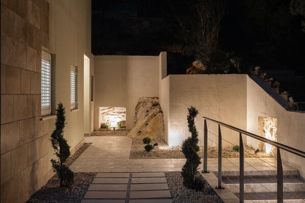 A nighttime view of a building's entrance with a stone pathway leading to a set of stairs, flanked by small trees and a metal railing.