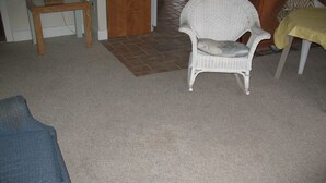 Una habitación con alfombra color beige, una silla de mimbre blanca, una mesa de madera y un sofá azul.