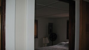 Una habitación tenuemente iluminada con una cama, un ventilador de pared y una mesita con una lámpara.