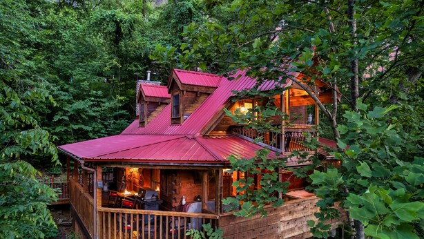 A wooden cabin with a red roof, surrounded by dense forest. It features a deck with outdoor seating and a fireplace. - Rustic Escape: Nature Views, Hot Tub & Fire Pit (Sevierville)