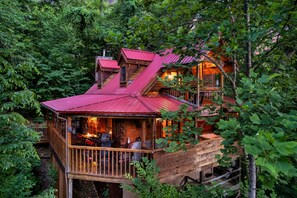A wooden cabin with a red roof, surrounded by dense forest. It features a deck with outdoor seating and a fireplace.