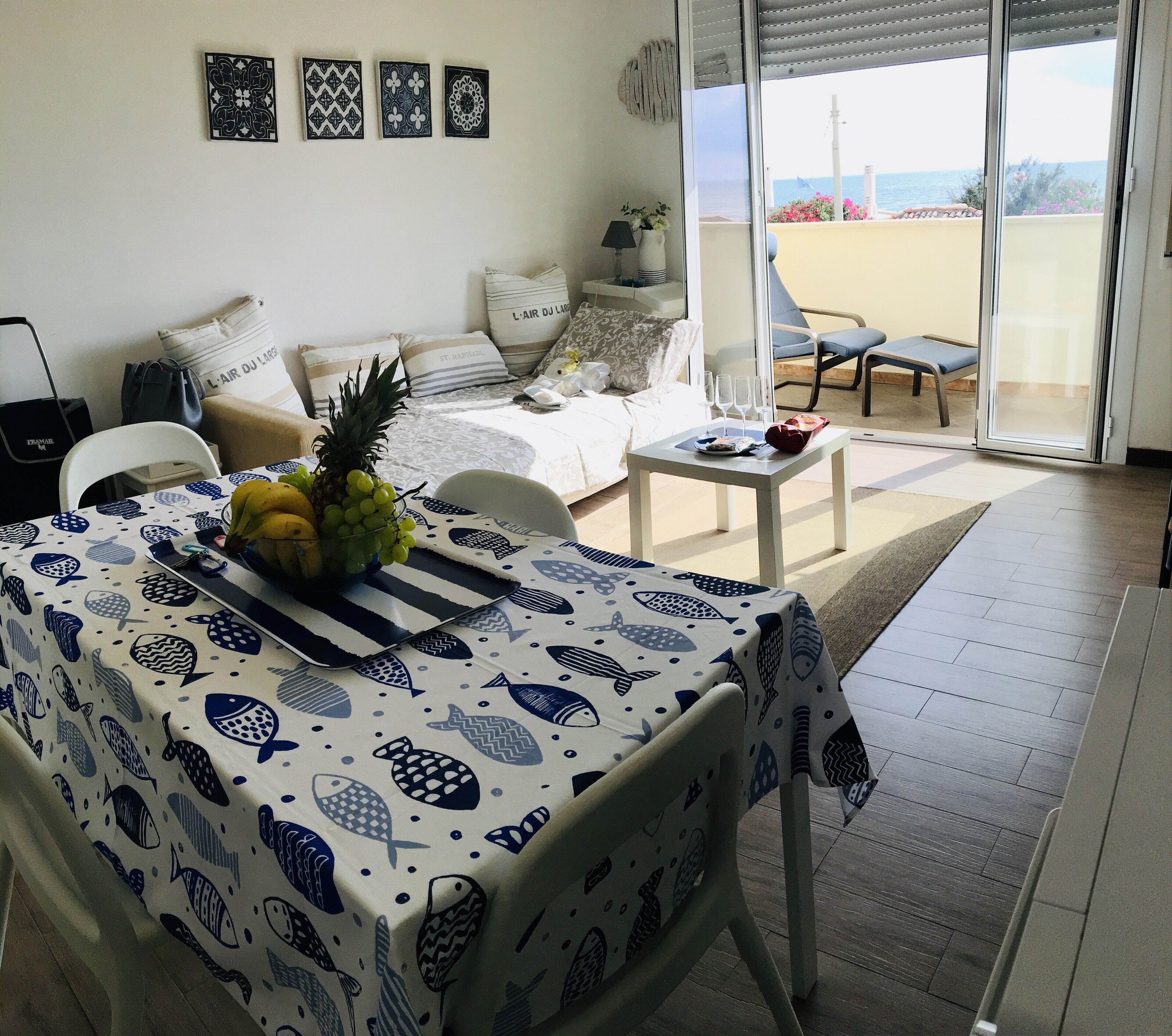 A dining area with a table covered in a fish pattern tablecloth, a pineapple, and grapes. There is a sofa with a striped pillow and a chair with a white cushion. A small table with a red bag and a glass of water is visible.