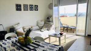 A dining area with a table covered in a fish pattern tablecloth, a pineapple, and grapes. There is a sofa with a striped pillow and a chair with a white cushion. A small table with a red bag and a glass of water is visible. - House facing the sea (Passoscuro)