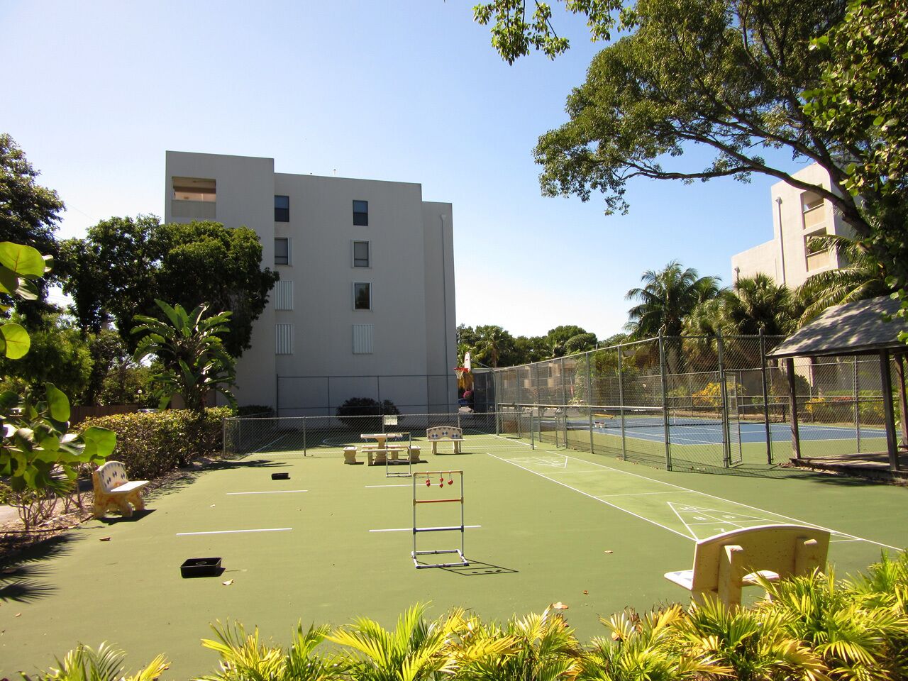 An outdoor tennis court with a fence, benches, and a building in the background.