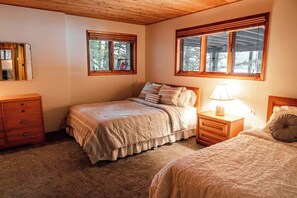 A bedroom with two beds, wooden headboards, a nightstand with a lamp, a dresser, and a window with a view of trees.