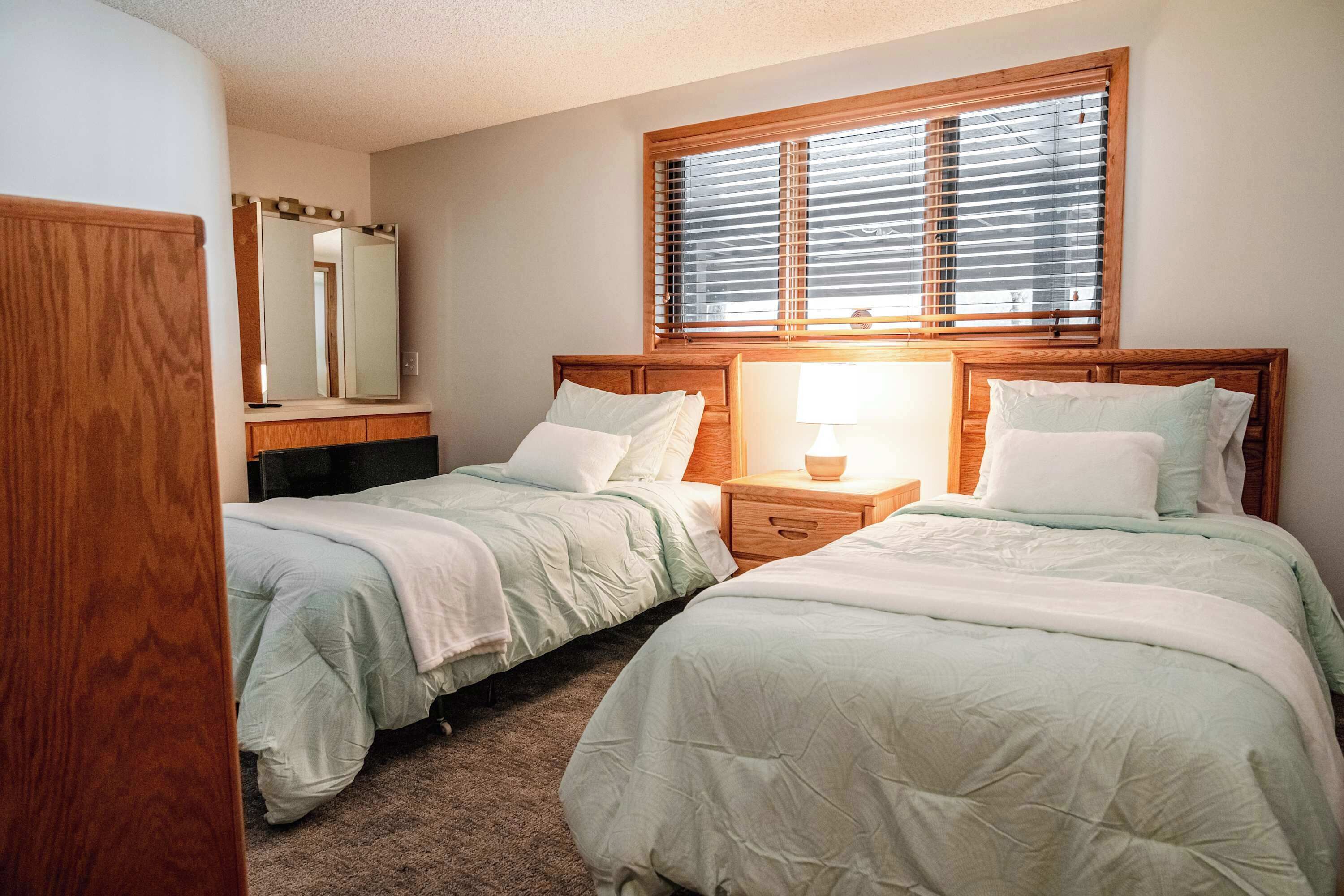 A bedroom with two beds, a nightstand, a mirror, and a window with blinds.