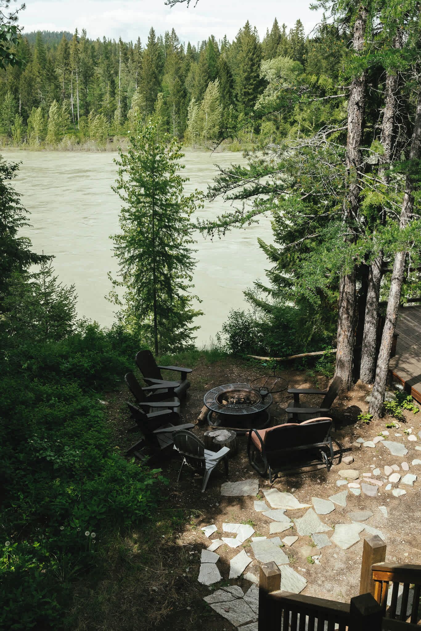 Un terrain de camping équipé d’un foyer, de chaises et offrant une vue sur une rivière bordée d’arbres.