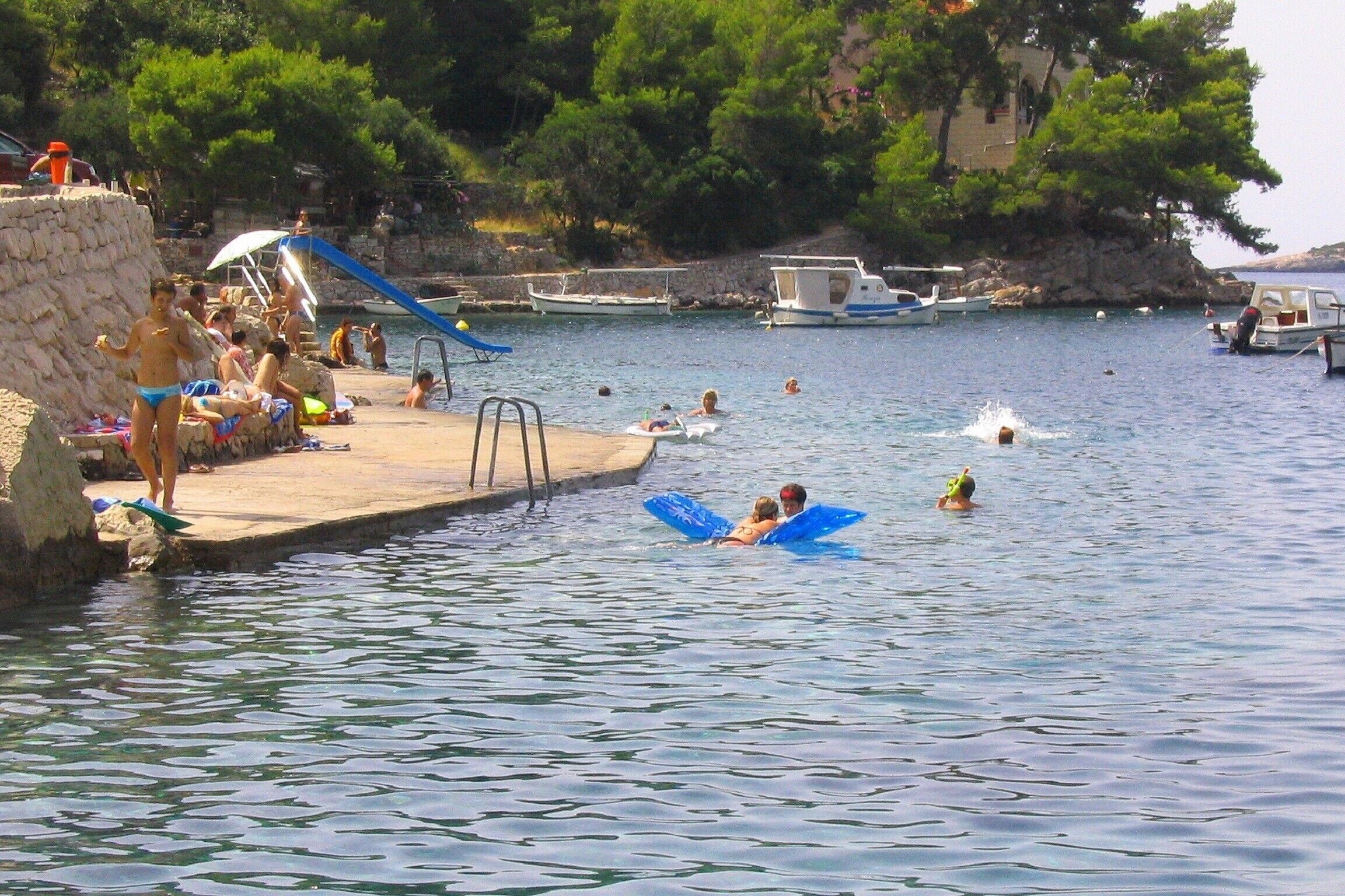 A beach with people swimming and sunbathing, boats in the water, and a stone wall.