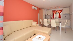 A modern living room with a beige sofa, a dining area with white chairs, and a kitchen with red curtains. - Apartments Korčula - Two Bedroom Apartment with Terrace and Sea View (Prižba)