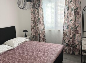 Un dormitorio con cama, mesita de noche, lámpara, cortinas con motivos florales y una ventana con persianas.
