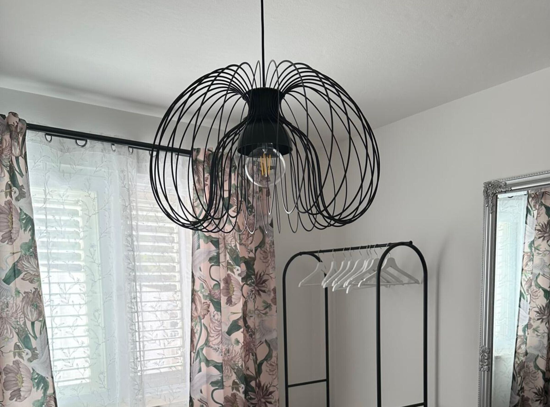 Um quarto com uma luminária de fio preto, cortinas florais, um suporte de metal e um espelho.