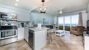 A modern kitchen with white cabinets, a central island, and stainless steel appliances. There is a dining area with a table and chairs, and a large window offering a view of the ocean.