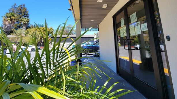 A covered outdoor area with a modern design, featuring a pathway, potted plants, and a view of parked cars and trees in the background. - Signature Inn Santa Clara (Santa Clara)