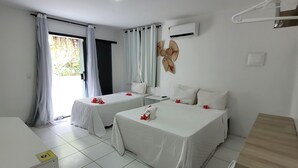 A hotel room with two beds, a wooden desk, an air conditioning unit, and a view of greenery outside. - Pousada Beach Garden (Ipojuca)