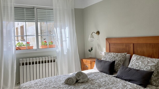 Une chambre à coucher avec un lit, une table de chevet, une lampe, une fenêtre avec des rideaux et un radiateur.