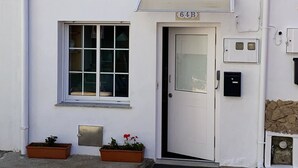 Un bâtiment blanc avec une porte portant l’inscription « 64B » et deux fenêtres. Des plantes en pot sont placées devant les fenêtres.
