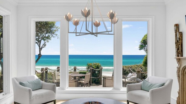 A room with a large window overlooking a beach, two white armchairs, a round coffee table, and a chandelier. - PoppyLu - Secluded Beachfront Home in Blue Mountain Beach with Private Pool & Golf Cart (Santa Rosa Beach)
