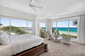 Un dormitorio con una cama grande, un ventilador de techo, vista a la playa y una pequeña mesa con sillas.