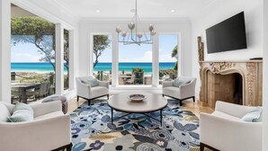 A spacious living room with a large window overlooking a beach, a fireplace, and a flat-screen TV mounted on the wall. - PoppyLu - Secluded Beachfront Home in Blue Mountain Beach with Private Pool & Golf Cart (Santa Rosa Beach)