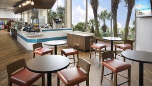 A restaurant with round tables and chairs, a counter with a display of fruits, and large windows with a view of palm trees and a beach.