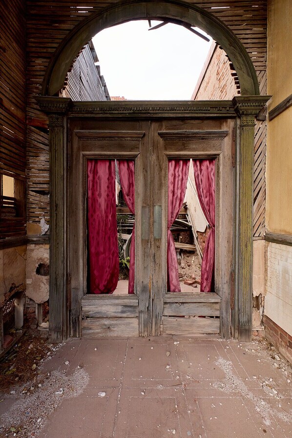 Un arco de entrada con paneles de madera y cortinas rojas, que conduce a un patio con escombros y un muro de ladrillo.