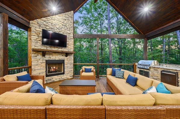 A covered outdoor seating area with stone walls, a flat-screen TV, a fireplace, and a grill.