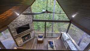 A cabin with a stone fireplace, a ceiling fan, a deck with outdoor seating, and a view of a forest. - Lux Cabin Private Pond Cust Playset 1 Mile to Lake Cvrd Deck+Entmt Amenities (Broken Bow)