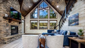 A spacious living room with a stone fireplace, large windows, a wooden ceiling, and a blue sofa. - Lux Cabin Private Pond Cust Playset 1 Mile to Lake Cvrd Deck+Entmt Amenities (Broken Bow)
