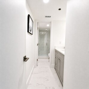 Un couloir étroit avec une porte de douche en verre, un lavabo blanc et un sol en marbre.