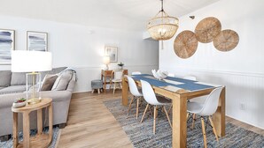 Una sala de estar moderna con comedor, que incluye una mesa de madera, sillas blancas, un sofá gris y una lámpara colgante de mimbre.