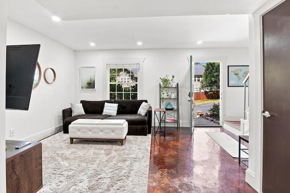 Un salon moderne avec un canapé marron foncé, un pouf blanc et un tapis à longues mèches blanc. Une fenêtre offre une vue sur une maison et des arbres, et une porte donne accès à l’extérieur.