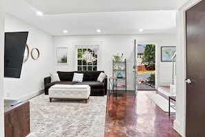 A modern living room with a dark brown sofa, a white ottoman, and a white shag rug. There is a window with a view of a house and trees, and a door leading outside.