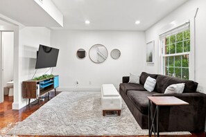 A modern living room with a flat-screen TV, a brown sofa, a white ottoman, and a large window with a view of greenery.