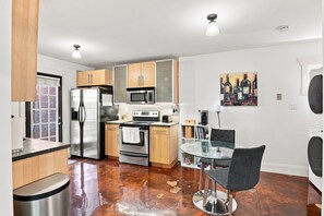 A modern kitchen with stainless steel appliances, wooden cabinets, a glass dining table, and a painting on the wall.